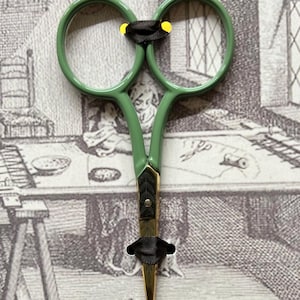 May include: Green handled embroidery scissors with gold blades and black accents. The scissors have a decorative design on the handle and are designed for precision cutting. The scissors are shown against a backdrop of an antique print.