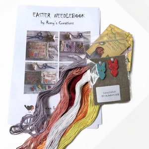May include: A needlepoint kit with instructions, embroidery floss, and two small ceramic bunny figurines. The kit is titled "Easter Needlebook" by Romy's Creations. The bunnies are red and blue.