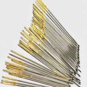 May include: A collection of sewing needles with gold-colored eyes and silver shafts, arranged in a fan shape against a white background. The needles are sharp and slender, designed for detailed stitching and craft projects.