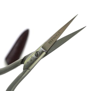 May include: Stainless steel scissors with pointed blades. The word "STAINLESS" is printed on one blade. The scissors have a silver handle and a small screw in the center. A dark brown object is visible in the background.