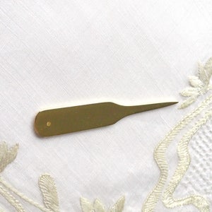 May include: A gold-colored metal tool with a pointed tip and a small hole near the handle. The tool is lying on a white surface.