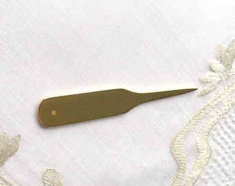May include: A gold-colored metal tool with a pointed tip and a small hole near the handle. The tool is lying on a white surface.