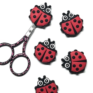May include: A pair of metal scissors with a pink and black leopard print pattern on the handles and blades, accompanied by four small, circular, red and black ladybug-shaped embellishments with white eyes and black pupils.