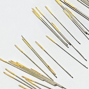 May include: A collection of sewing needles with gold-colored eyes and silver shafts, scattered on a white surface. The needles are of varying lengths and are designed for hand sewing and crafting projects.