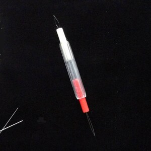 May include: A clear plastic needle threader with a red and white tip. The threader has two needles attached.