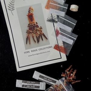 Peut inclure: Un kit d'artisanat pour créer un masque perlé, comprenant un patron imprimé, de petits sachets de perles de différentes couleurs, des aiguilles et du fil. Le patron est intitulé "THE MASK" et comprend le logo Fern Ridge Collections.