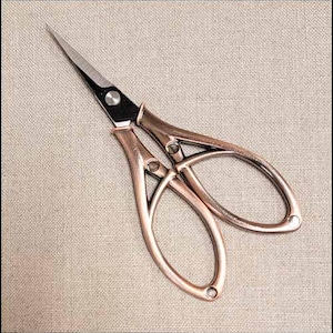 May include: A pair of small, copper-colored scissors with a pointed blade. The scissors have a decorative design on the handles and a black section near the pivot point. The scissors are designed for detailed cutting tasks.