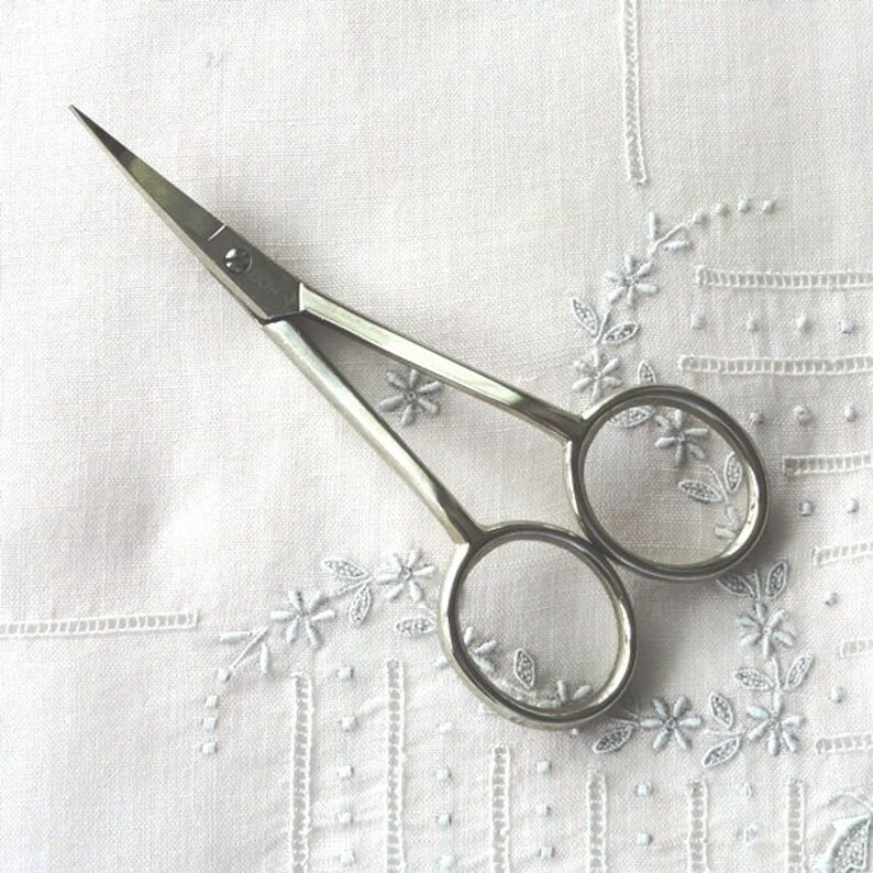 Puede incluir: Tijeras de bordado plateadas con una punta afilada y dos anillos ovalados. Las tijeras est&aacute;n sobre una tela blanca con un dise&ntilde;o bordado.