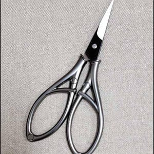 May include: A pair of silver-toned embroidery scissors with a sharp, pointed blade. The scissors have a unique, open-loop handle design and are set against a neutral, textured background. The blades are black and silver.