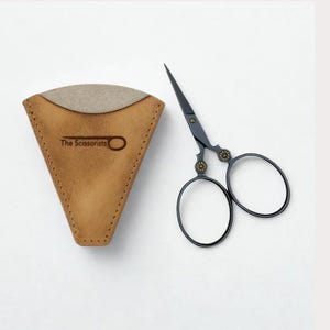 May include: A pair of black scissors with decorative gold accents and a tan leather case. The case is shaped like a triangle with the text "The Scissorists" printed on it. The scissors are designed for precision cutting.