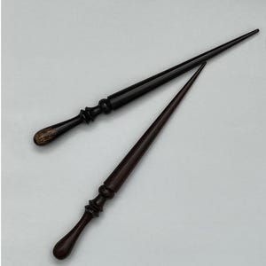 Könnte beinhalten: Zwei dunkelbraune Holzhaarnadeln. Ein Stift hat ein abgerundetes Ende und der andere ein spitzes Ende.