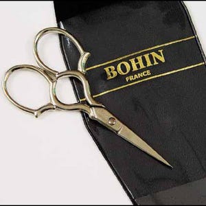 May include: Silver-toned Bohin France embroidery scissors with curved handles and sharp blades, resting on a black leather case. The case features gold lettering that reads "BOHIN FRANCE". The scissors are designed for precision cutting.