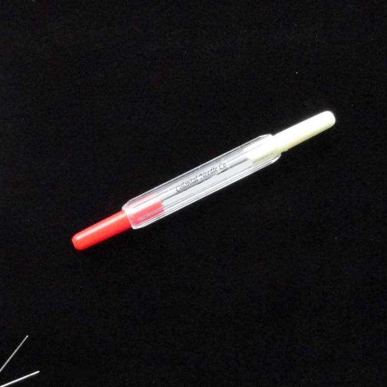 May include: A clear plastic needle threader with a red tip on one end and a white tip on the other end. The threader has the text "Colonial Syringe Co." printed on the side.