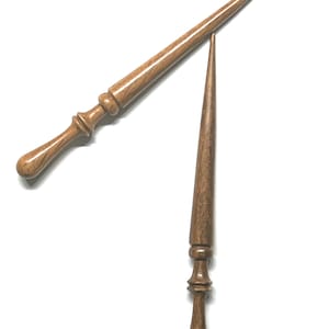 May include: Two hand-turned wooden knitting needles with a smooth, polished finish and a natural brown wood grain. Each needle features a tapered point at one end and a decorative, rounded handle with carved rings at the base of the shaft.