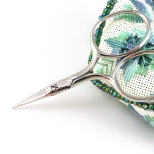 May include: Silver embroidery scissors with a pointed tip and a small screw on the handle. The scissors are resting on a green and white fabric with a beaded border.