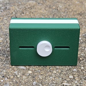 May include: A rectangular, dark green box with a white stripe along the top edge. A white, circular knob is centered on the front, with two horizontal slots. The box is sitting on a textured, gray surface.