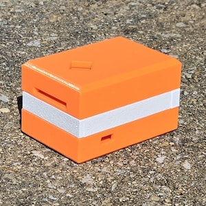 May include: Bright orange rectangular object with a white stripe. The object has a slot on the front and a small rectangular opening on top. The object is sitting on a rough, gray surface.