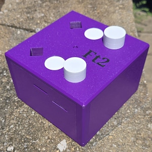 May include: A purple box with the text "Ft2" printed on the top. The box has two slots and several white circular and cylindrical objects on top. The box is sitting on a rough, gray surface.