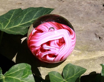 Könnte beinhalten: Ein Glas-Briefbeschwerer mit rosa und weißen Wirbeln im Inneren. Der Briefbeschwerer ist rund und hat eine klare Glasaußenseite.