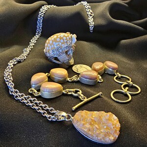Peut inclure: Un ensemble de bijoux comprenant un collier en chaîne argentée, une bague avec un motif blanc et jaune et un bracelet avec des pierres ovales. Un pendentif avec un grand cristal orange est également présent. Les bijoux sont présentés sur un fond sombre.