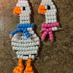 May include: Two handmade beaded keychains shaped like geese, constructed from white, orange, blue, and pink plastic pony beads. The larger goose features a blue scarf, while the smaller goose has a pink scarf, both with gold-tone metal rings.