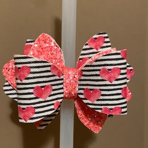 May include: A pink and black hair bow with a white stripe pattern and pink hearts. The bow is made of fabric and has a glittery finish.