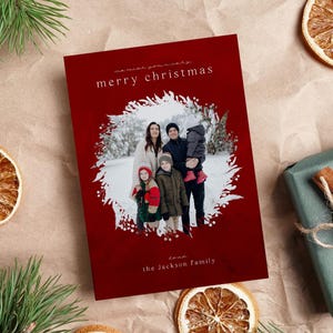 May include: A rectangular Christmas greeting card with a deep red background featuring a central circular photo of a family of four in a snowy setting. The card includes the white text merry christmas and the jackson family in a minimalist font.