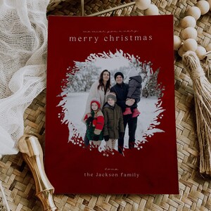 May include: A rectangular Christmas card with a deep red background featuring a central circular photo of a family in a snowy landscape framed by a white botanical wreath. The text reads we wish you a very merry christmas and love the Jackson family.