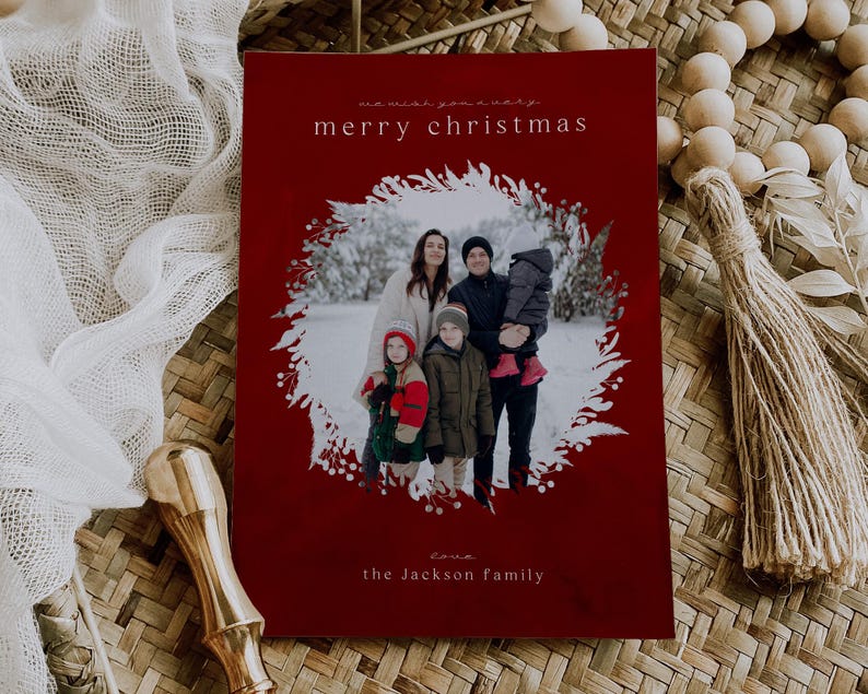 May include: A rectangular Christmas card with a deep red background featuring a central circular photo of a family in a snowy landscape framed by a white botanical wreath. The text reads we wish you a very merry christmas and love the Jackson family.