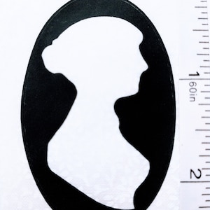 May include: A black and white cameo silhouette of a woman's profile, set within an oval frame. The image shows the woman's hair styled in a bun. The cameo is approximately 5 cm in height.