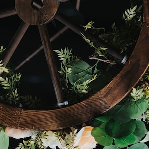 Könnte beinhalten: Ein Wagenradkranz aus Holz, verziert mit künstlichem Grün und pfirsichfarbenen Blüten. Der Kranz ist mit einem kleinen Metallschild verziert, auf dem "Vintique" steht.