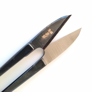 May include: A close-up of thread snips, with black handles and silver blades. The blades are open, showing a sharp edge. A small gold inscription is visible on the black handle. These snips are designed for precision cutting of threads and small materials.