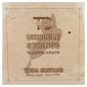 May include: A square package of Toda Guitars fluorocarbon ukulele strings with a textured tan paper finish. The front features the TD logo, the text UKULELE STRINGS, FLUOROCARBON, TODA GUITARS, Shiga Japan, and Since 1998, framed by a thin border.
