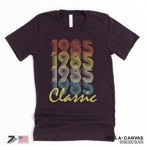 Pu&ograve; includere: Una maglietta marrone scuro con un design grafico in stile vintage che presenta l'anno 1985 ripetuto quattro volte in uno schema di colori arcobaleno. Il testo "Classic" &egrave; stampato sotto i numeri in un carattere giallo.