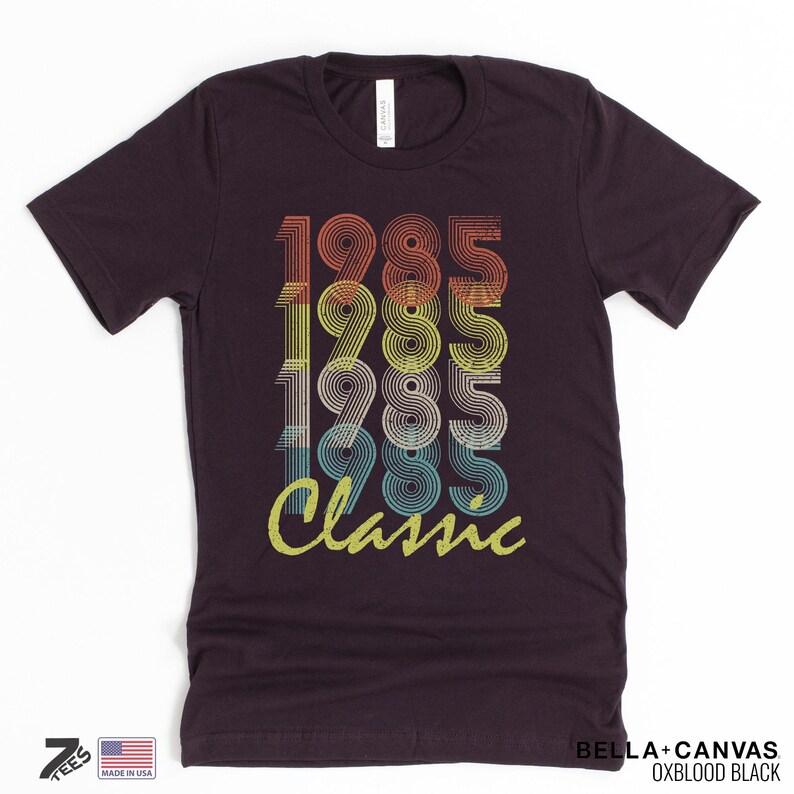 Pu&ograve; includere: Una maglietta marrone scuro con un design grafico in stile vintage che presenta l'anno 1985 ripetuto quattro volte in uno schema di colori arcobaleno. Il testo "Classic" &egrave; stampato sotto i numeri in un carattere giallo.