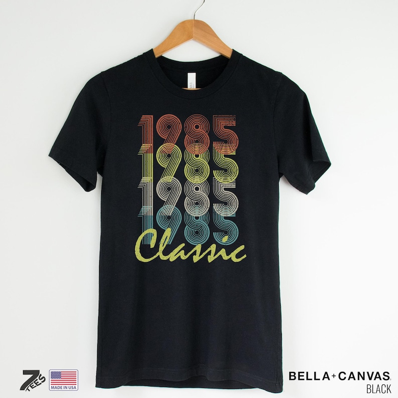 Pu&ograve; includere: Maglietta nera con una grafica in stile vintage che dice "1985 Classic" in colori arcobaleno.