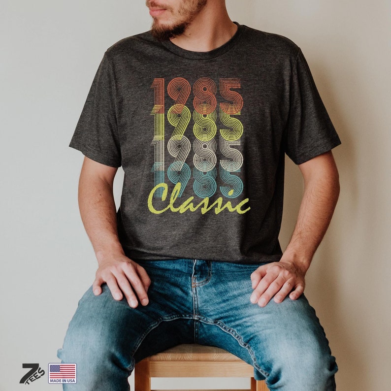 Pu&ograve; includere: Una maglietta grigio scuro con un design grafico in stile vintage che presenta l'anno 1985 ripetuto quattro volte in un arcobaleno di colori, con la parola "Classico" sotto.