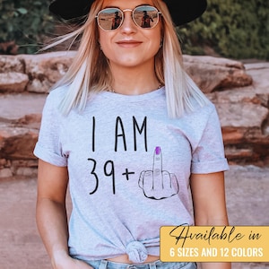 Pode incluir: Uma camiseta cinza com o texto "I AM 39+" e um emoji de dedo do meio com esmalte de unha roxo.