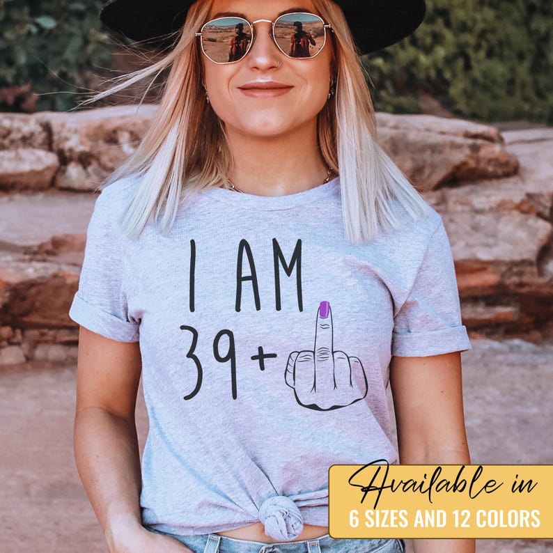 Pode incluir: Uma camiseta cinza com o texto "I AM 39+" e um emoji de dedo do meio com esmalte de unha roxo.