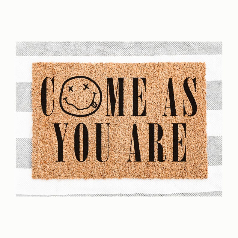 May include: A rectangular doormat made of natural coir fiber. The mat features the phrase "COME AS YOU ARE" in bold black letters, with a smiley face graphic above the word "ME". The mat is set against a striped background.