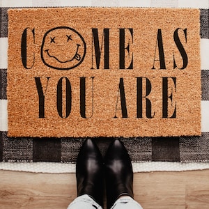 May include: A rectangular doormat made of natural coir fiber, featuring the phrase "COME AS YOU ARE" in black capital letters, along with a smiley face with crossed-out eyes and a protruding tongue. The mat is placed on a black and white checkered rug.