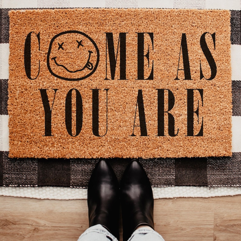 May include: A rectangular doormat made of natural coir fiber, featuring the phrase "COME AS YOU ARE" in black capital letters, along with a smiley face with crossed-out eyes and a protruding tongue. The mat is placed on a black and white checkered rug.