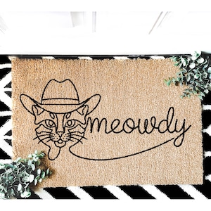 May include: A brown doormat with a black line drawing of a cat wearing a cowboy hat and the word "meowdy" written in a cursive font.