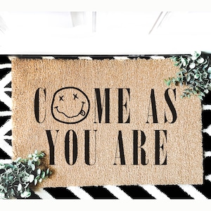May include: A rectangular doormat with the words "COME AS YOU ARE" in black block letters. The word "COME" features a smiley face with crossed-out eyes and a tongue sticking out. The mat is tan with a black and white patterned border.