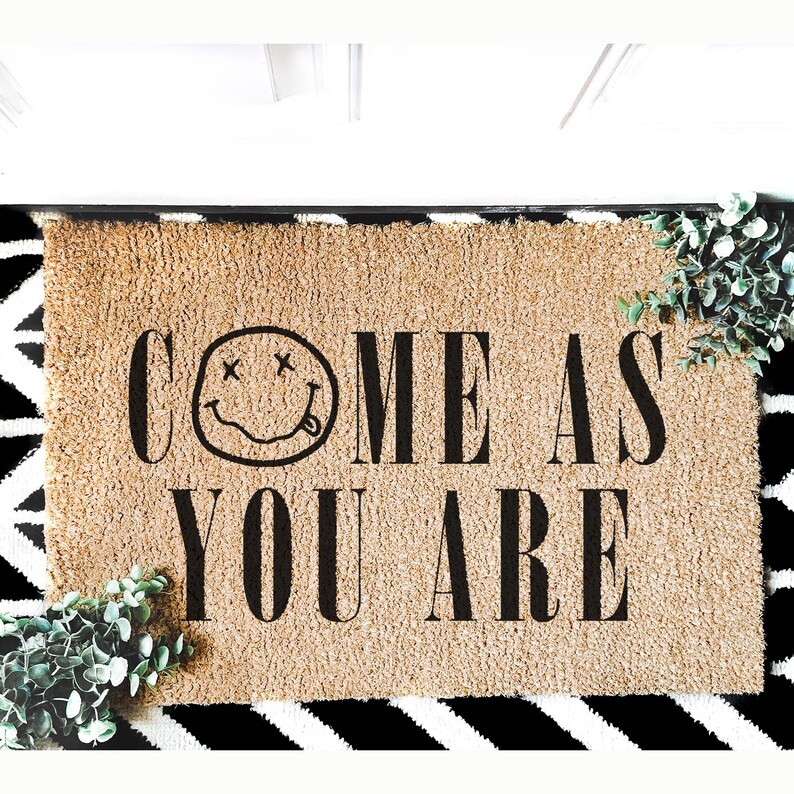 May include: A rectangular doormat with the words "COME AS YOU ARE" in black block letters. The word "COME" features a smiley face with crossed-out eyes and a tongue sticking out. The mat is tan with a black and white patterned border.