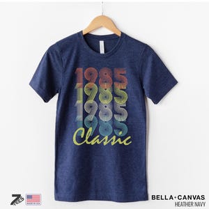 Pu&ograve; includere: Una t-shirt blu navy con una grafica in stile vintage che dice "1985 Classic" in colori arcobaleno.