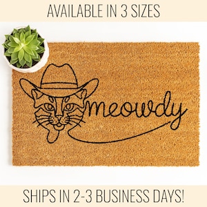 May include: A brown coir doormat with a black line drawing of a cat wearing a cowboy hat and the word "meowdy" written in a cursive font.
