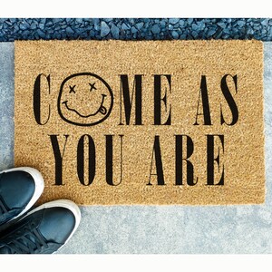 May include: A rectangular doormat made of natural coir fiber. The mat features the phrase "COME AS YOU ARE" in bold black letters, with a smiley face graphic. Black sneakers are visible at the bottom left corner of the mat.