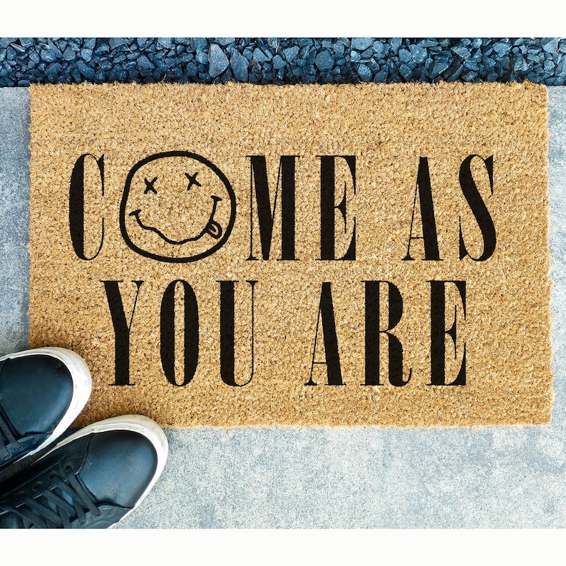 May include: A rectangular doormat made of natural coir fiber. The mat features the phrase "COME AS YOU ARE" in bold black letters, with a smiley face graphic. Black sneakers are visible at the bottom left corner of the mat.