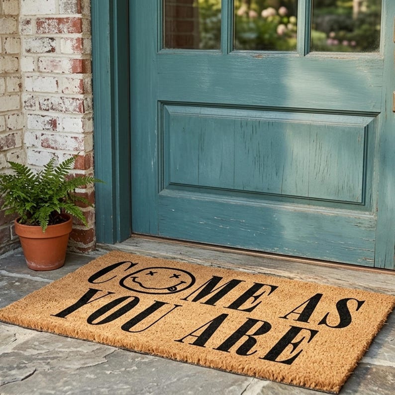 May include: A rectangular coir doormat with the words "COME AS YOU ARE" in black, along with a smiley face design. The mat is placed in front of a teal door and a brick wall, with a potted fern to the left.